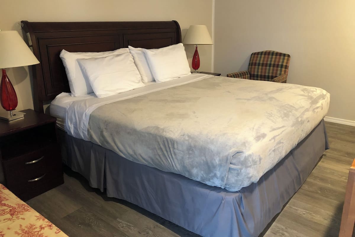 A neatly made bed with a headboard, two bedside lamps, a plaid chair, and a wooden floor.