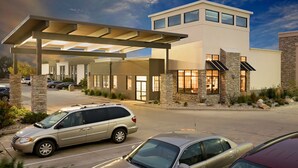 Un edificio moderno con acceso cubierto, fachada de piedra y ladrillo, y un estacionamiento con automóviles.