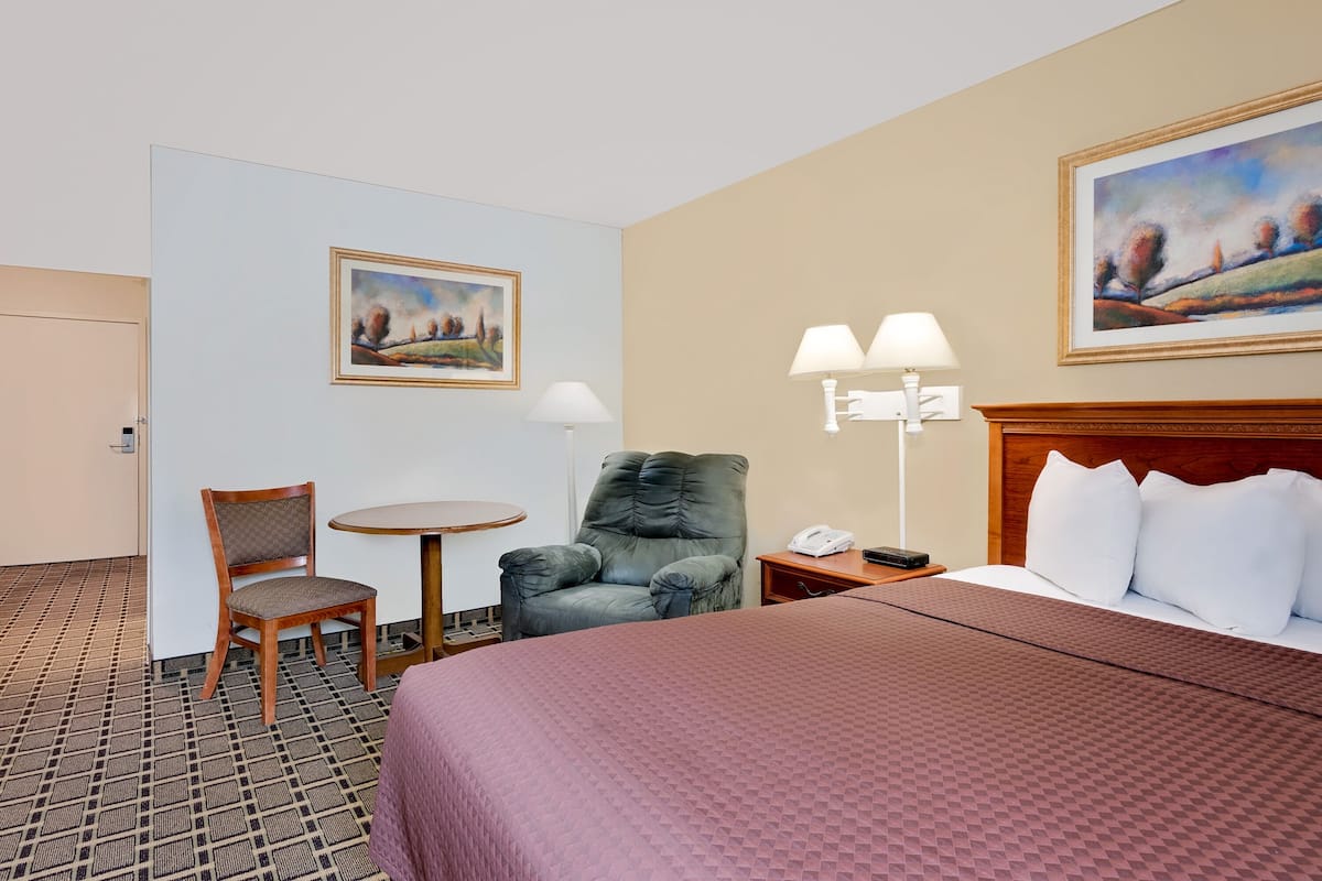 A hotel room with a bed, a chair, a table, and two paintings on the walls.