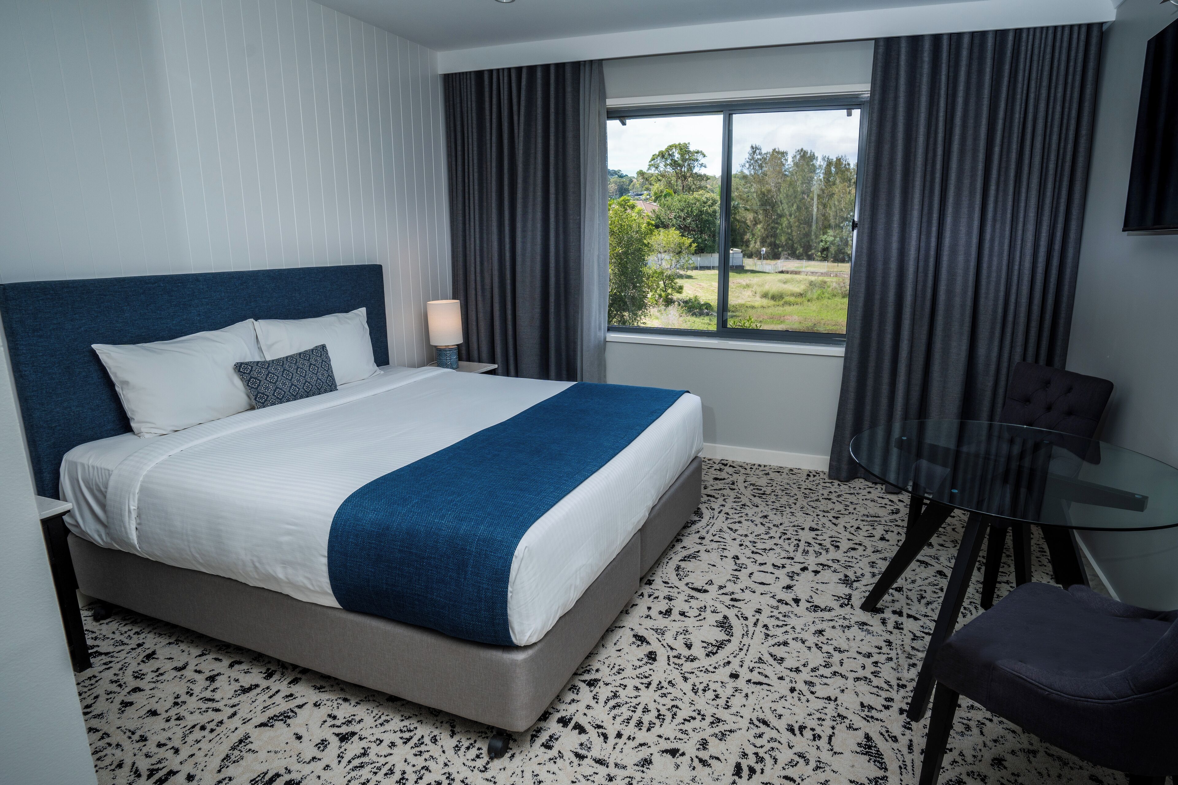 A hotel room with a large bed, a glass table, a chair, and a view of the outdoors.