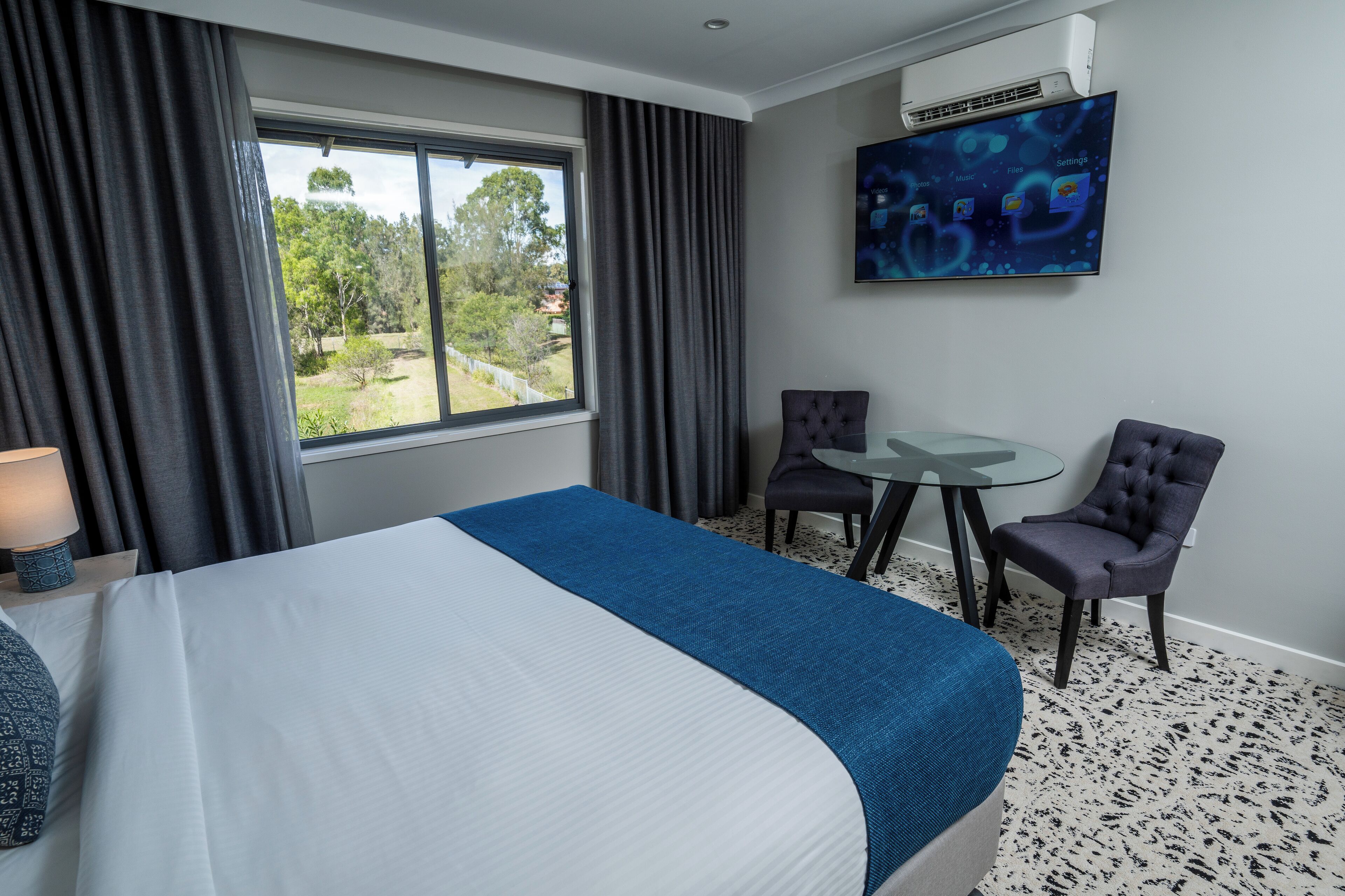 Habitación de hotel con una cama grande, un escritorio con dos sillas, un televisor en la pared y una ventana con cortinas.