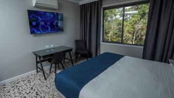 A hotel room with a bed, a desk, a chair, a TV mounted on the wall, and a window with curtains.