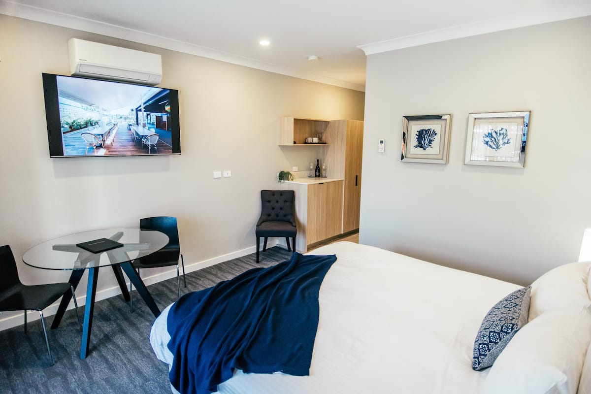 A hotel room with a bed, a desk with a chair, a small kitchenette, and a TV mounted on the wall.