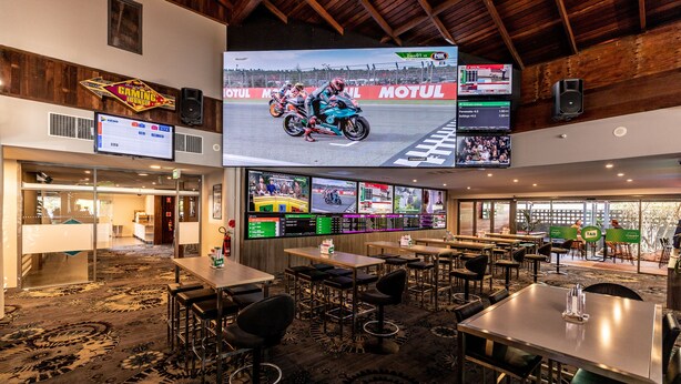 A hotel lobby with multiple screens displaying motorcycle racing, a bar area, and a seating area with tables and chairs. - Warners At The Bay (Warners Bay)