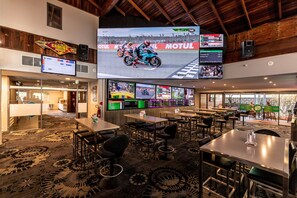 A hotel lobby with multiple screens displaying motorcycle racing, a bar area, and a seating area with tables and chairs. - Warners At The Bay (Warners Bay)