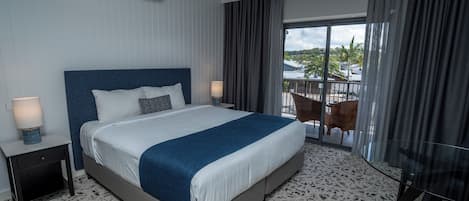A hotel room with a large bed, a nightstand with a lamp, a glass table, and a view of the outdoors through a sliding glass door.