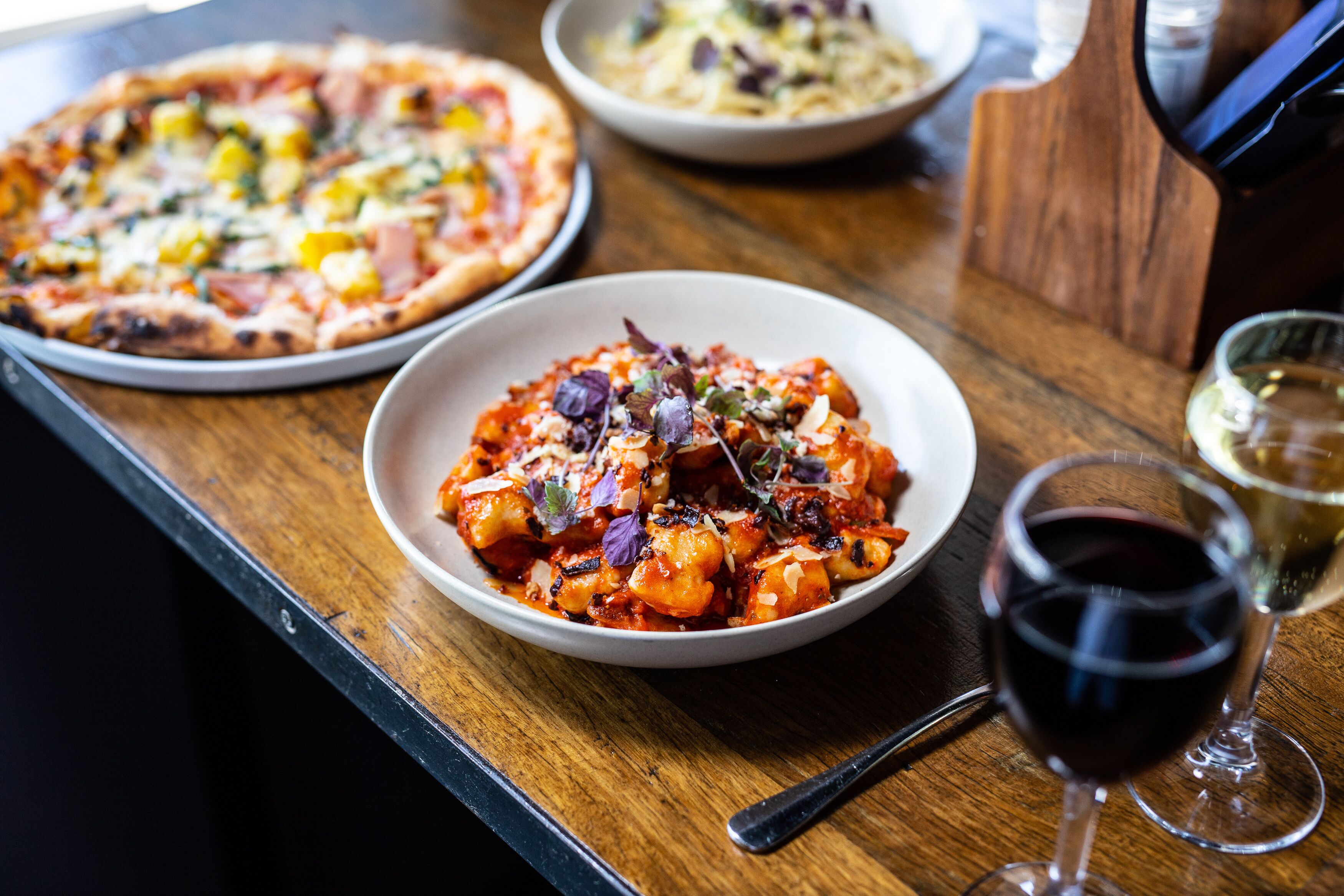 A wooden table with a pizza, a bowl of pasta, and a glass of red wine.