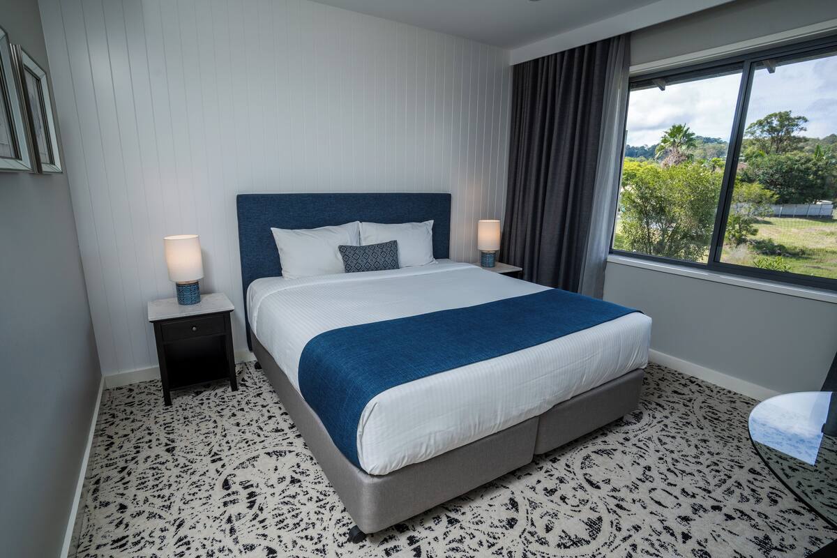 A hotel room with a large bed, two bedside tables, a window with a view of greenery, and a patterned carpet.