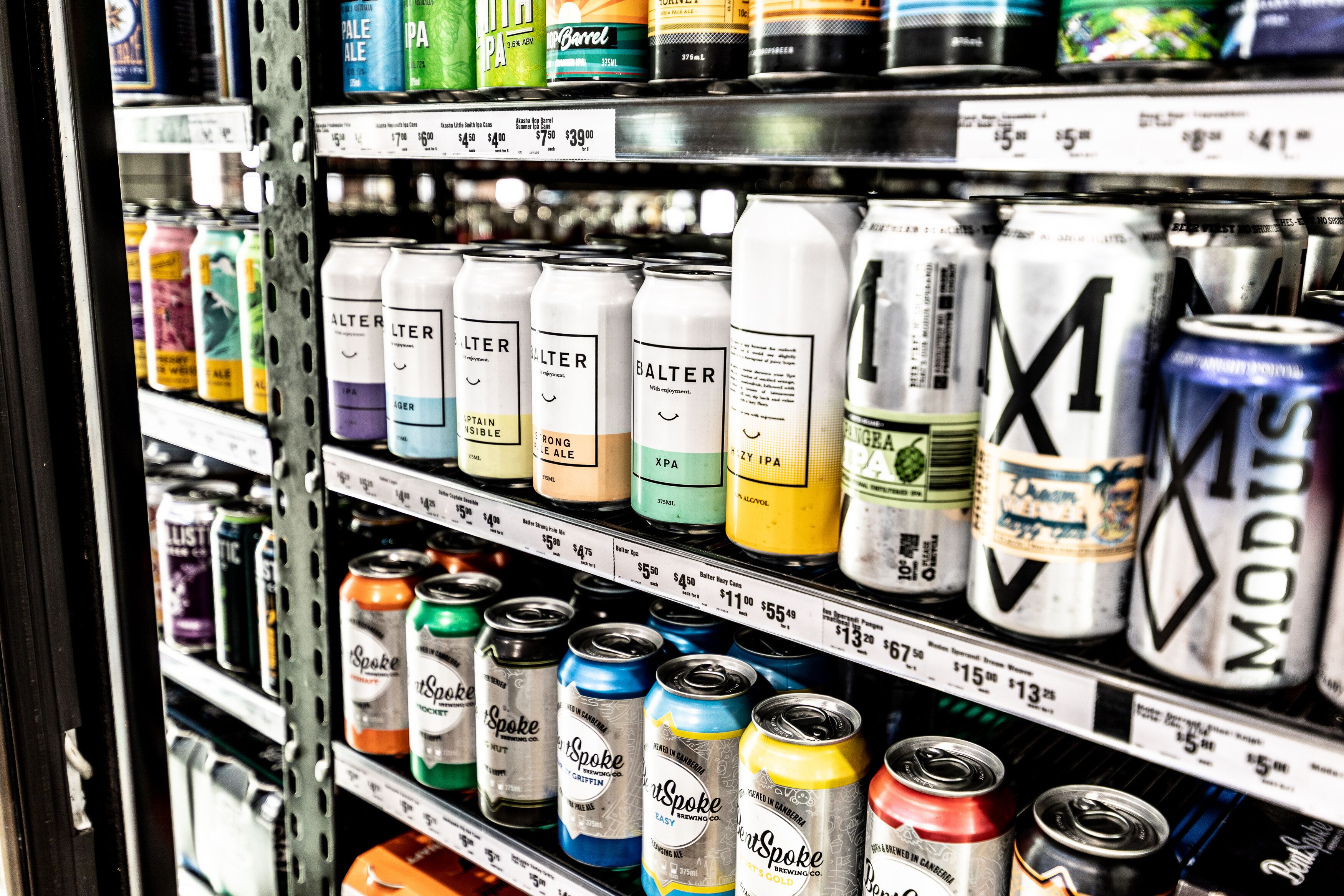 A shelf stocked with various beer cans and bottles, including brands like Balter and Spoke, with price tags visible.