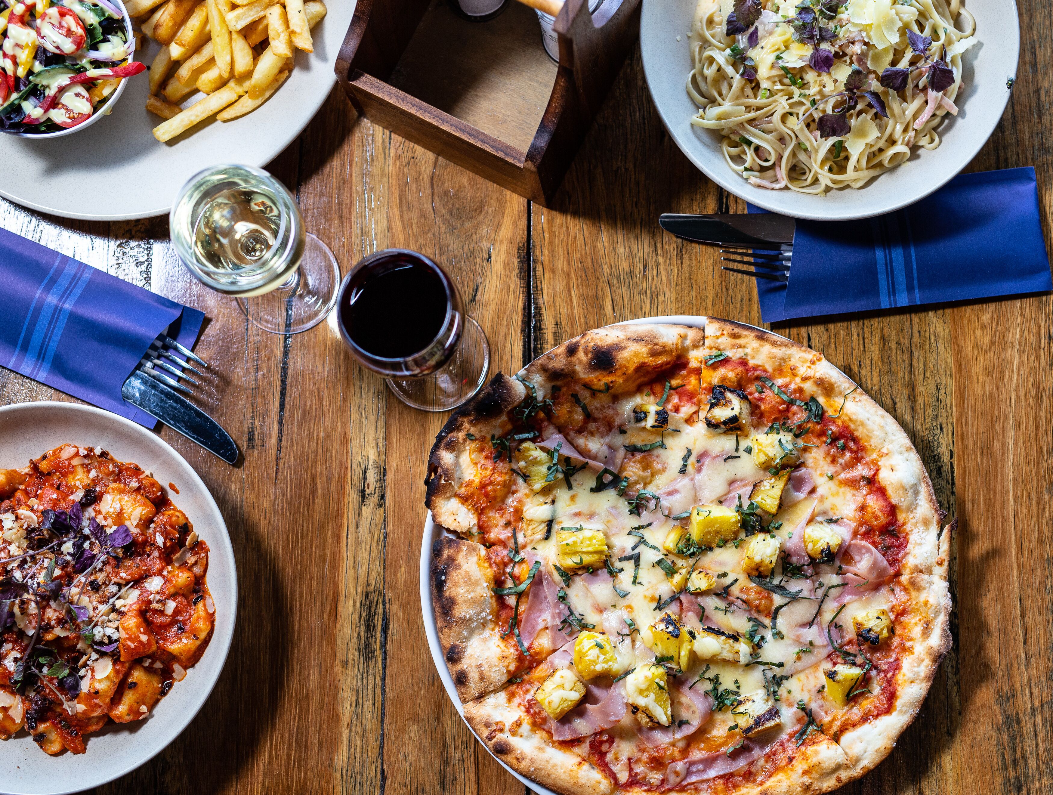 A wooden table with various dishes including a pizza, pasta, fries, and a salad.