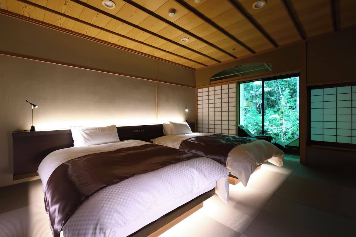 Un dormitorio de estilo japonés tradicional con suelo de tatami, un bajo cabecero de madera y vistas a la vegetación a través de un panel shoji.