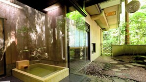 Un onsen japonès tradicional amb una banya de fusta, una font termal amb envàs de vidre i un camí de pedra que condueix a una porta de fusta.