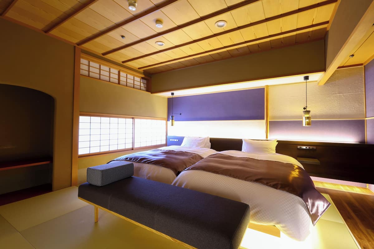Un dormitorio de estilo japonés tradicional con suelo de tatami, techo de madera bajo y una cama grande con muchos almohadones.