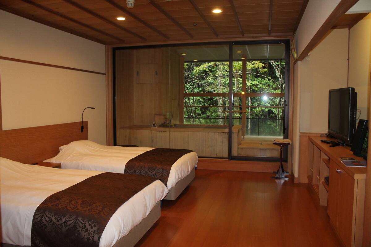 Habitación de hotel con dos camas, televisión y un amplio ventanal corredero que da a un balcón con vistas a árboles.
