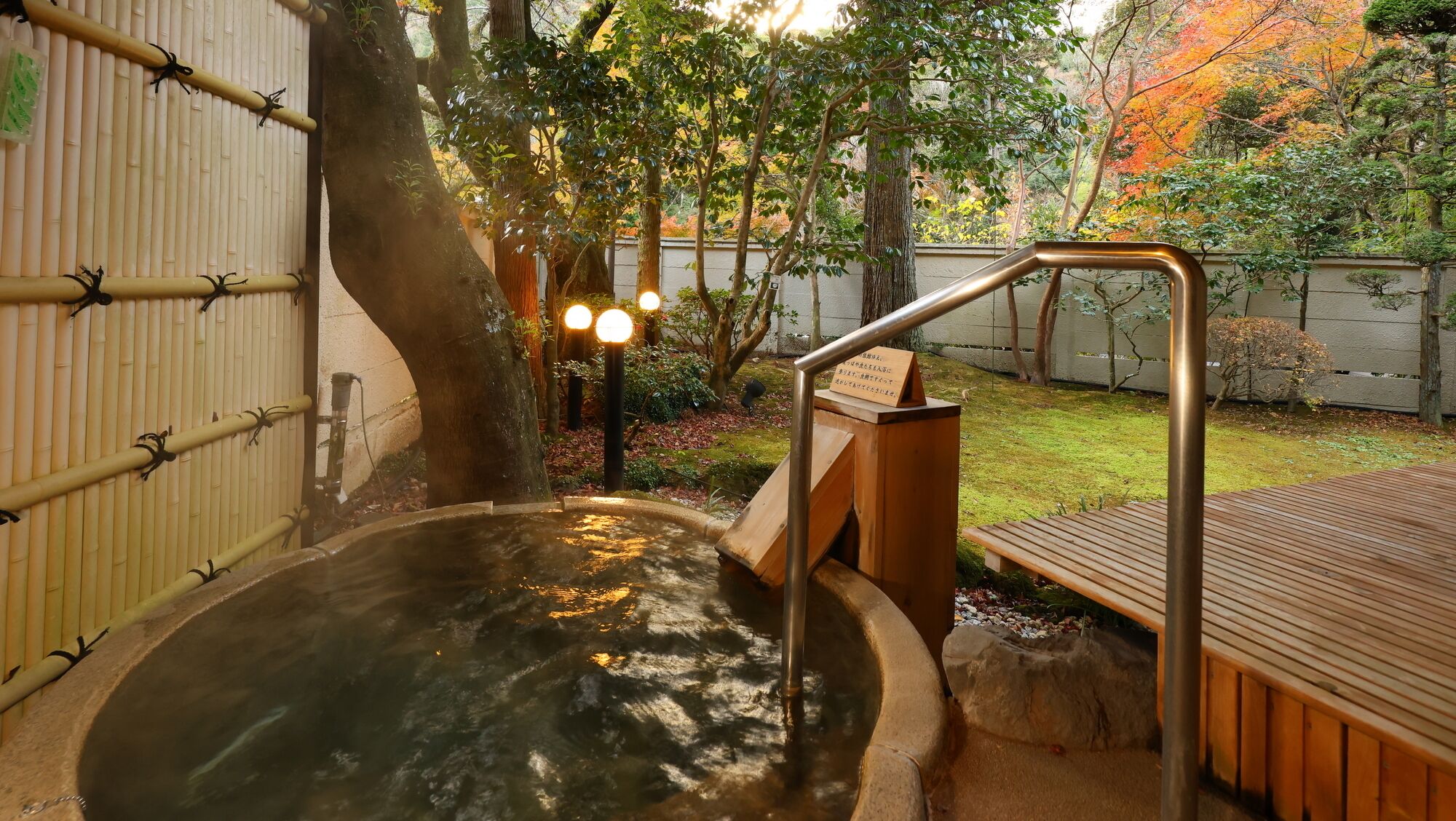 Zona de baño de aguas termales con una terraza de madera, valla de bambú y un sendero de piedra que conduce al baño.