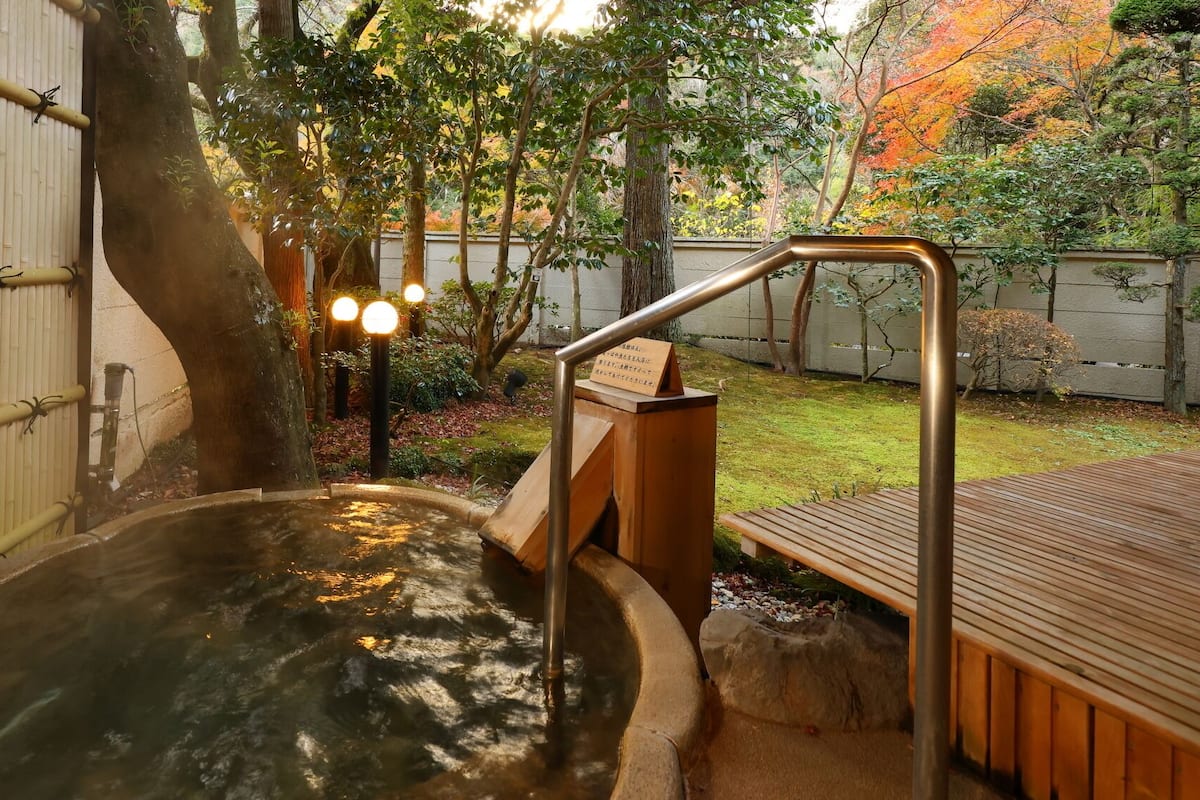 Zona de baño de aguas termales con una terraza de madera, valla de bambú y un sendero de piedra que conduce al baño.