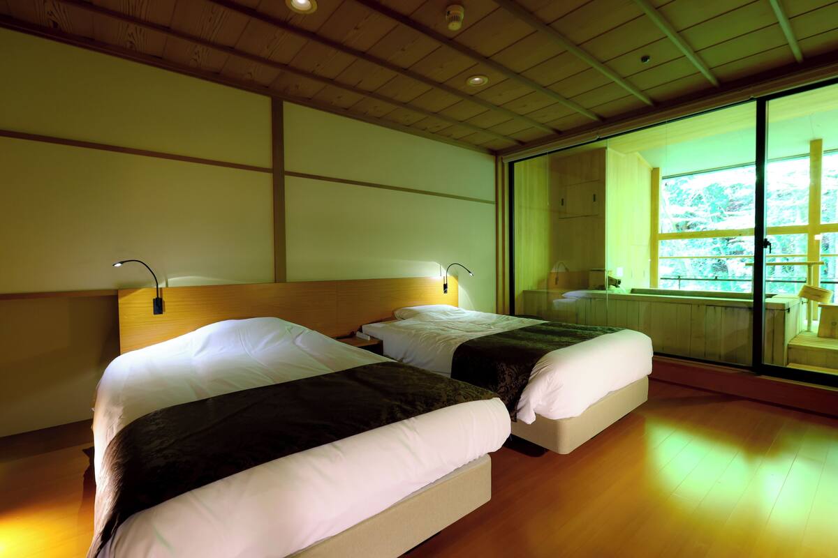 Habitación de hotel con dos camas, suelo de madera, un ventanal grande y vista a zonas verdes.