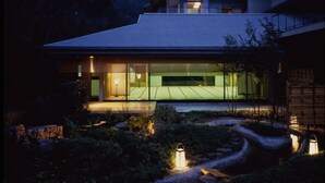 Una casa moderna amb una gran finestra de vidre, un interior ben il·luminat i un jardí amb camins i llums.