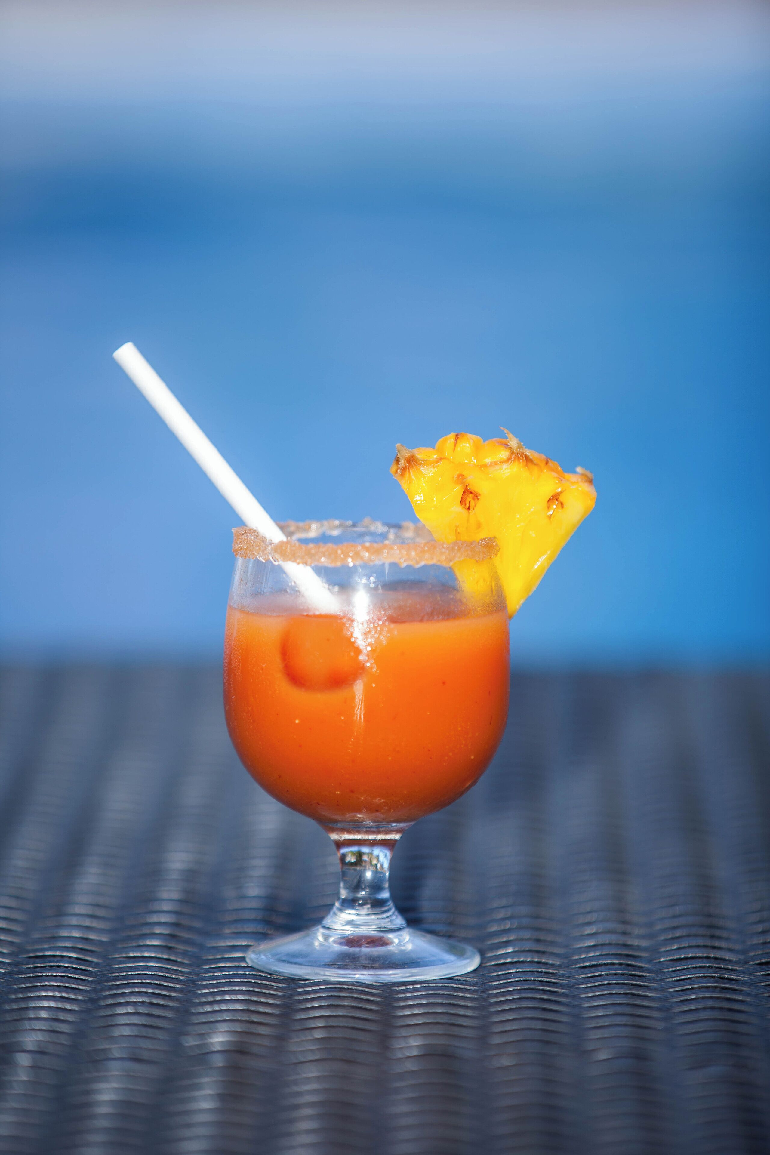 A cocktail glass with a slice of pineapple on the rim, a straw, and a textured surface beneath.