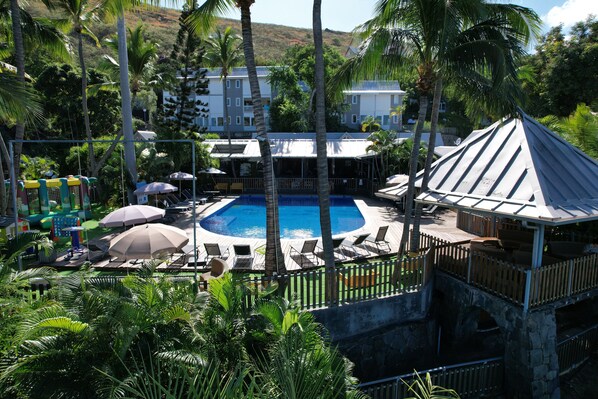 A pool area with lounge chairs and umbrellas, surrounded by palm trees and a stone bridge. - Les Aigrettes (Saint-Paul)