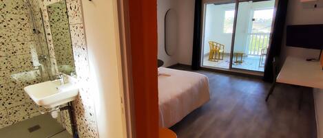 Habitación de hotel con baño, una cama, una mesita pequeña y un balcón con sillas.