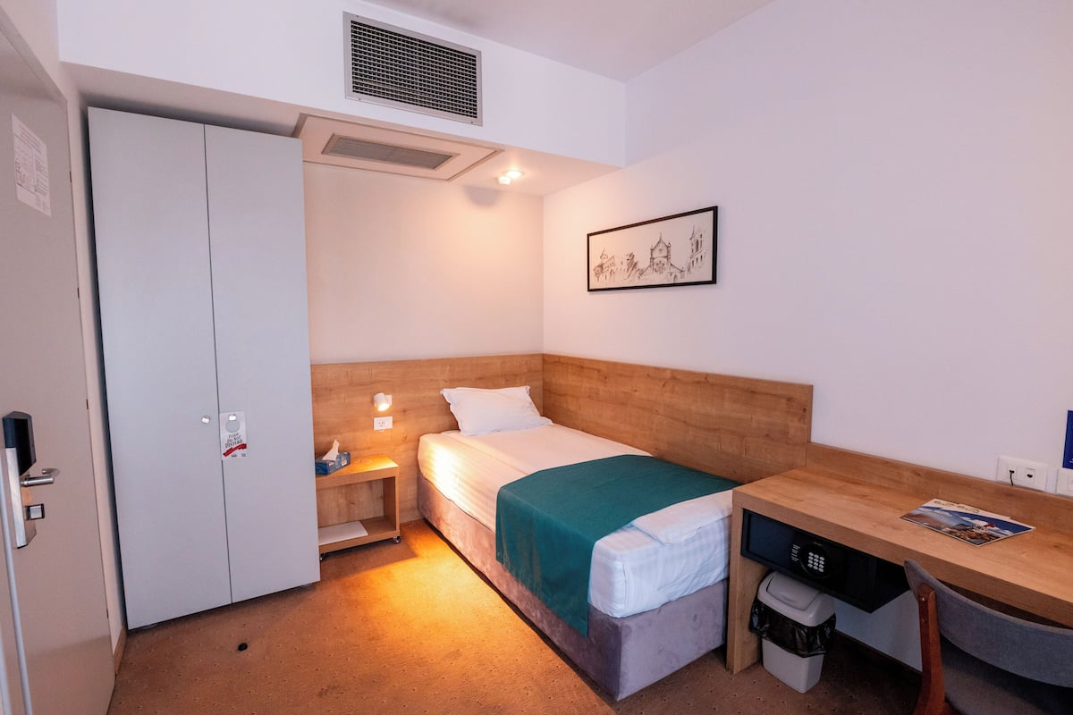A compact hotel room with a single bed, a desk, a chair, a wardrobe, and a small bedside table.
