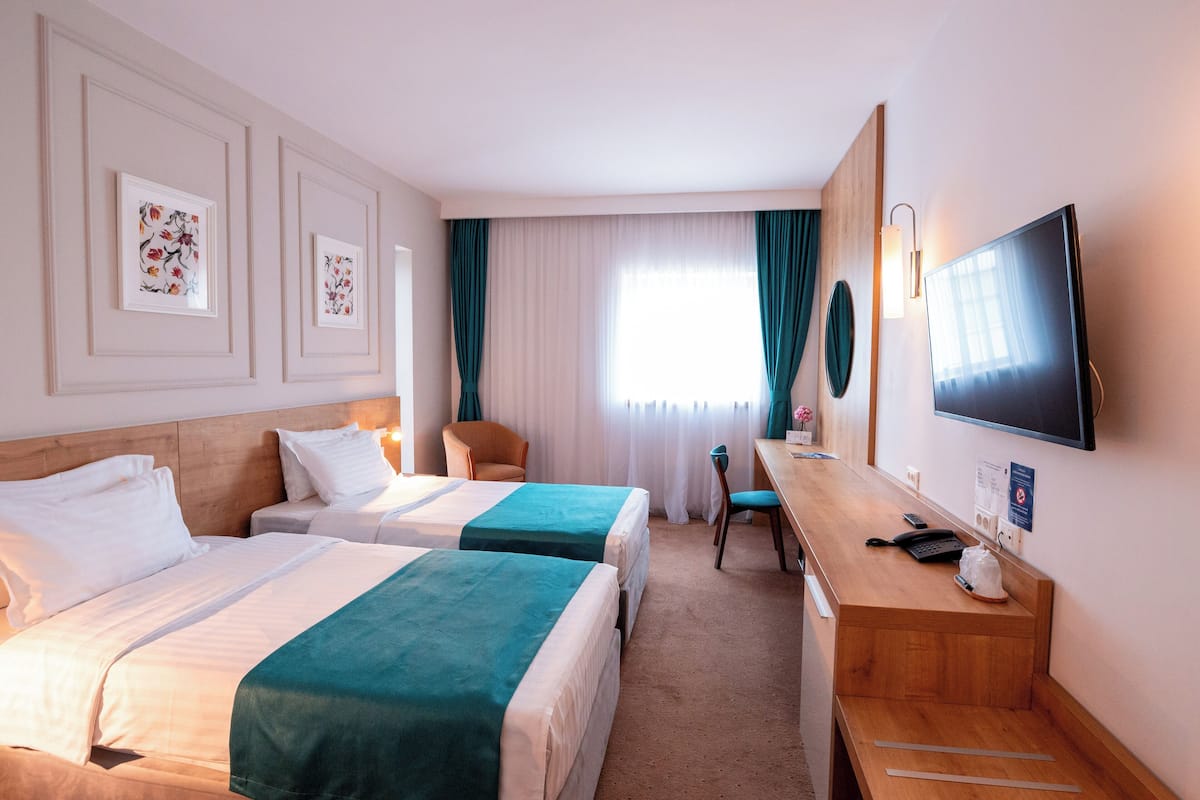 A hotel room with two beds, a desk, a TV, and a window with curtains.