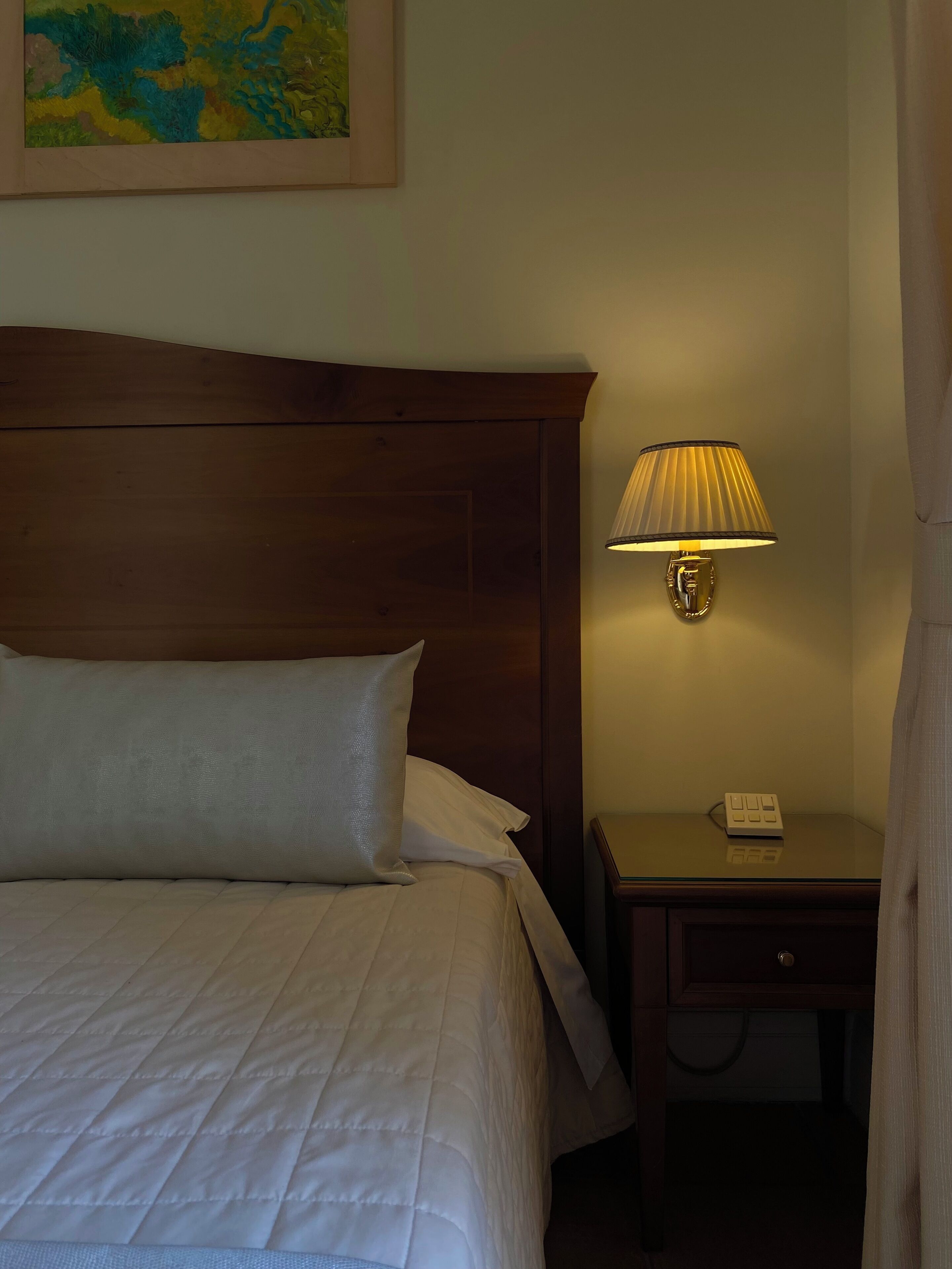 Una cama bien hecha con sábanas blancas, cabecera de madera, lámpara de noche y mesita con un teléfono.