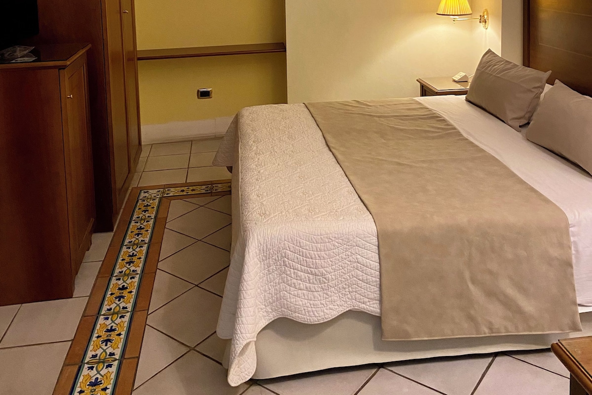 Habitación de hotel con cama, armario, estante con libros, lámpara y cuadros en la pared.