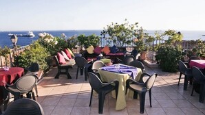 An outdoor patio with tables and chairs, a small seating area with cushions, and a view of the sea. - Akropolis Hotel (Taranto)
