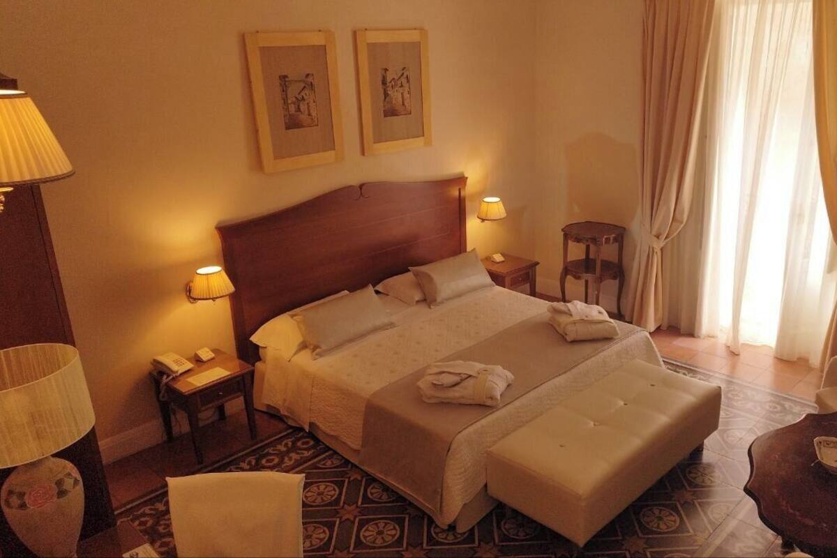 Habitación de hotel con una cama grande, dos lámparas de noche, una mesita, una silla y un televisor sobre un mueble.