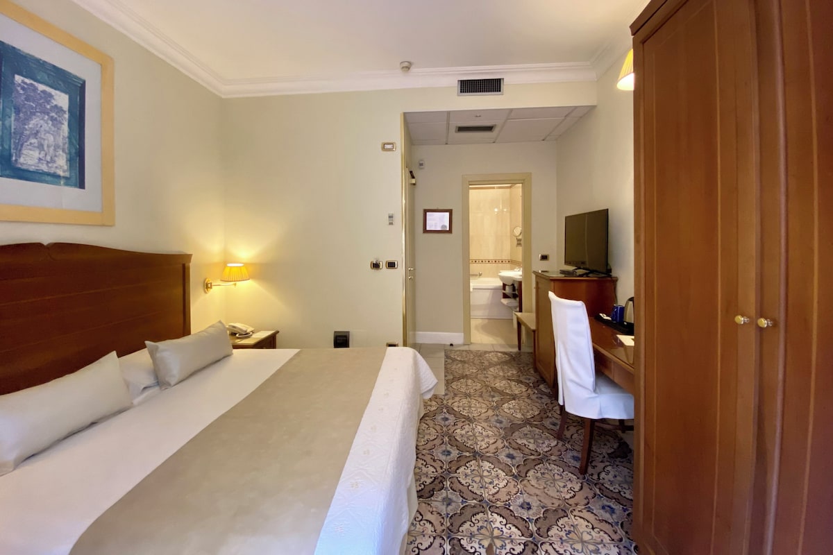 A hotel room with a large bed, bedside table, lamp, and a television.