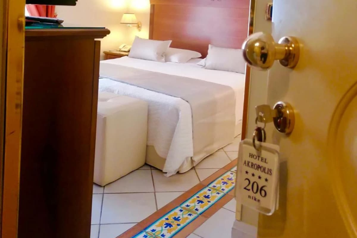 Habitación de hotel con una cama, una mesita de noche, una lámpara, un cuadro en la pared y una puerta con placa con el nombre del hotel.