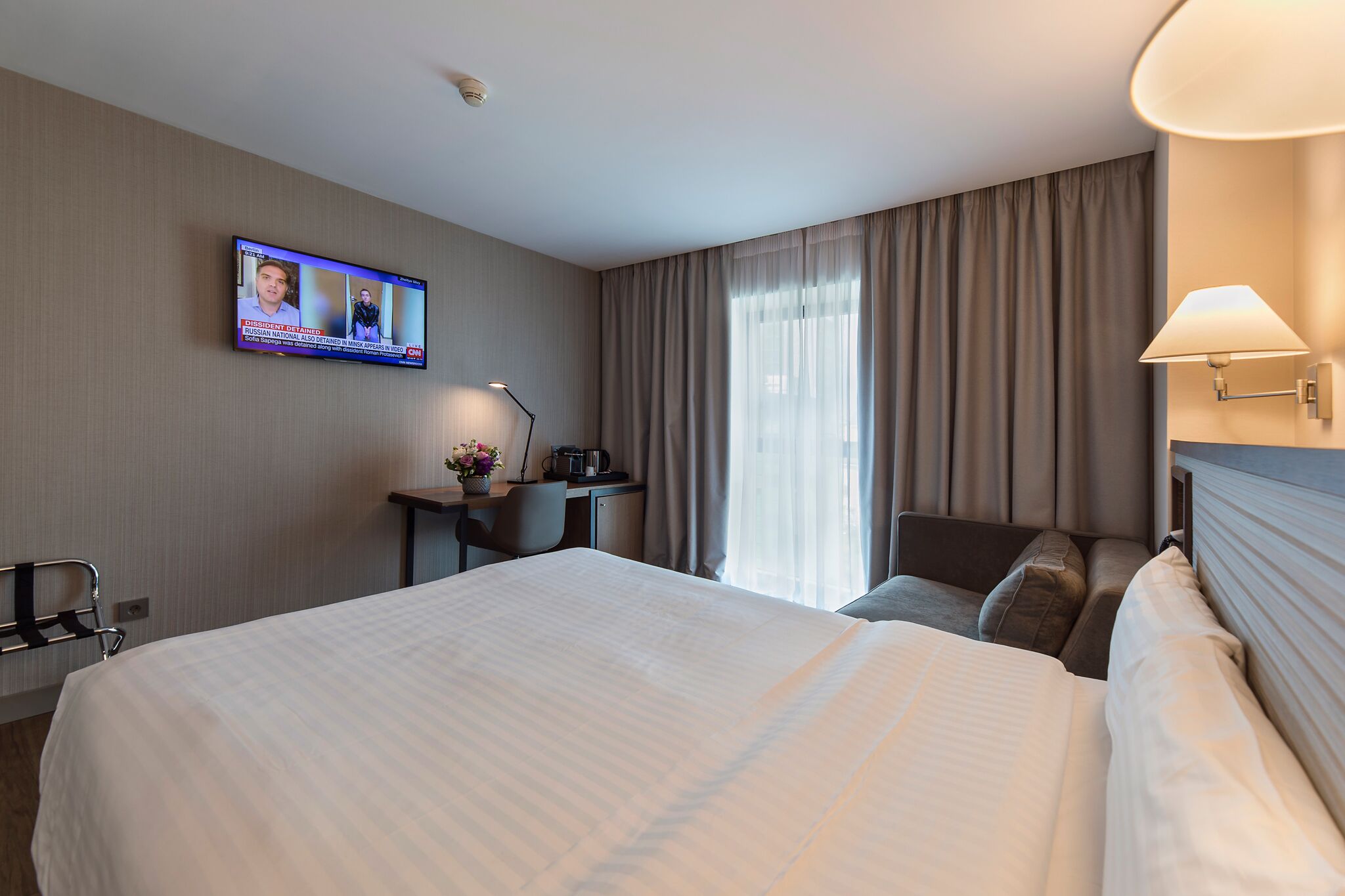 Habitació d'hotel amb un llit gran, una taula de treball amb una cadira, una televisió penjada a la paret i una finestra amb cortines.