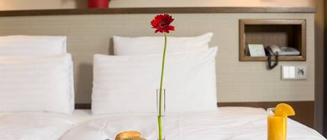 A neatly made bed with white linens, a tray with breakfast items including fruits, bread, and a glass of orange juice, and a red flower in a vase.
