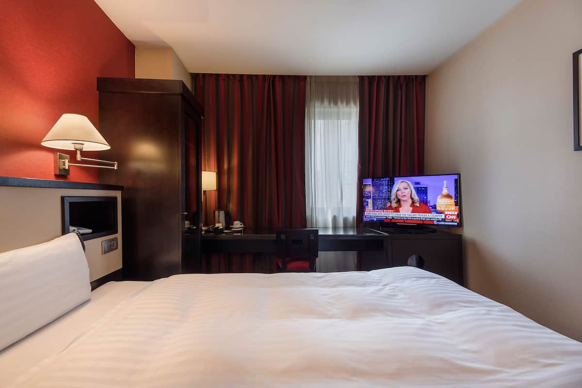Habitación de hotel con cama, escritorio con lámpara, televisor mostrando un noticiero y ventana con cortinas.