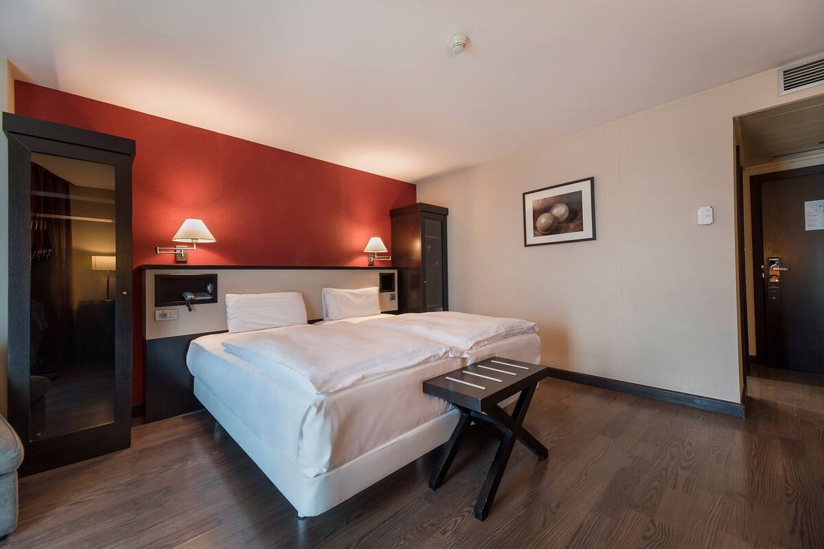 Una habitación de hotel con una cama, dos lámparas de noche, una mesita pequeña, un armario y una puerta.