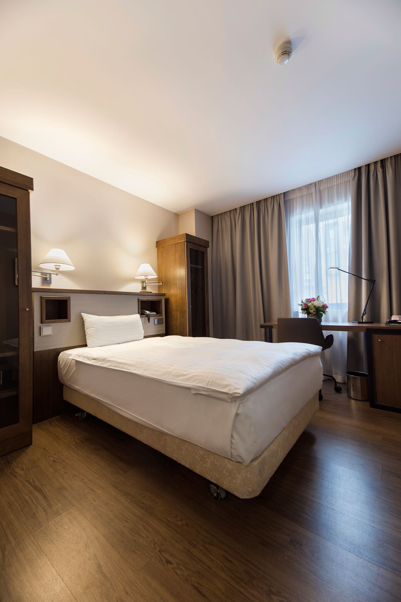 A hotel room with a large bed, a desk with a lamp, a wardrobe, and a window with curtains.