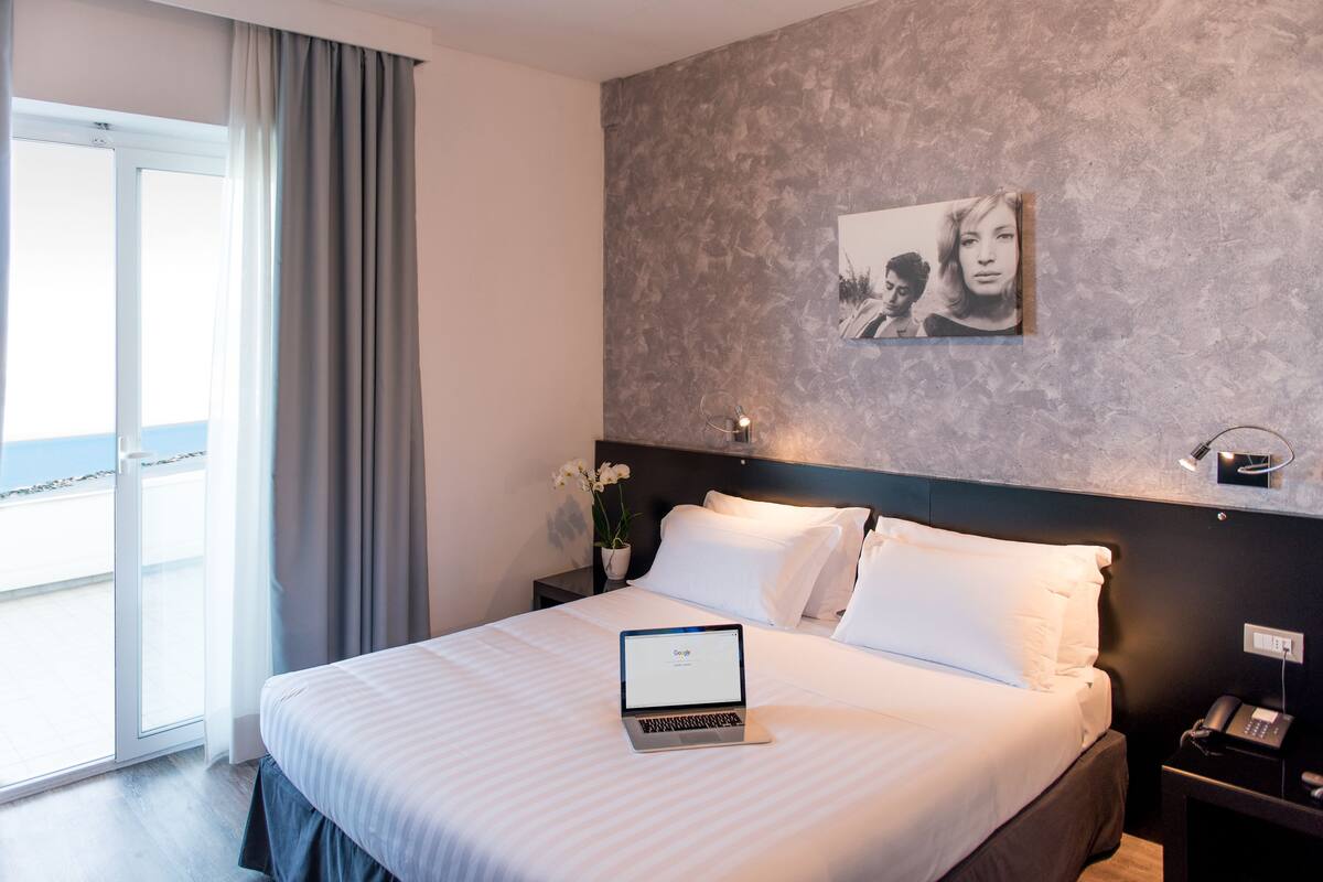 A hotel room with a bed, a laptop on the bed, a nightstand with a phone, a wall-mounted light, and a window with curtains.