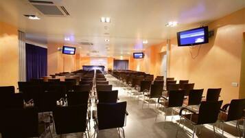 Una sala conferenze con file di sedie, un palco nella parte anteriore e due schermi alle pareti.