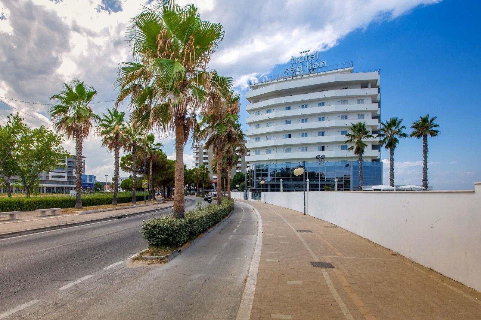 A modern hotel with a clear view of the sea, palm trees lining the street, and a well-maintained sidewalk.