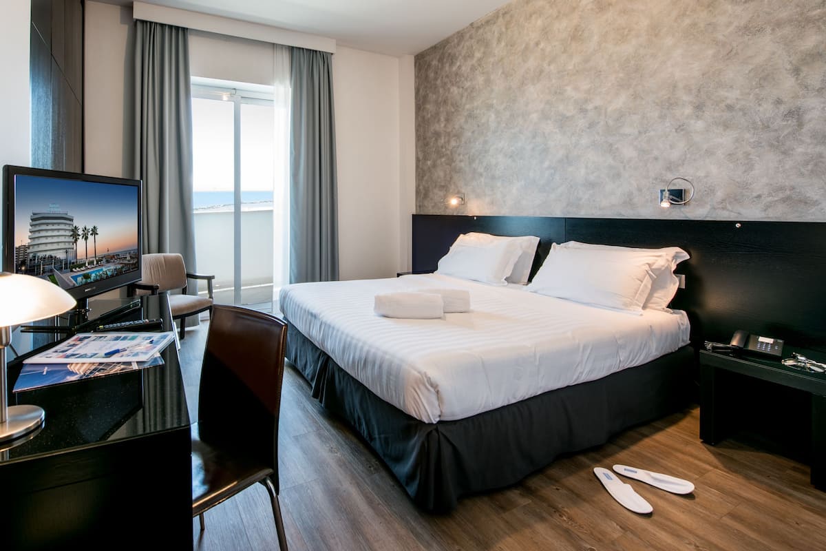 A hotel room with a bed, a desk with a computer, a chair, a TV displaying a cityscape, and a view of the beach through the window.