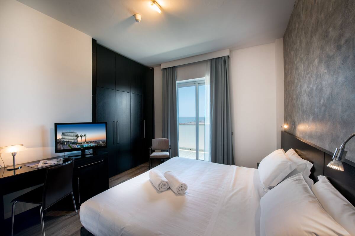 A hotel room with a bed, a desk, a television, and a view of the sea through a window.