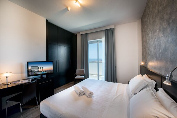 A hotel room with a bed, a desk, a television, and a view of the sea through a window.