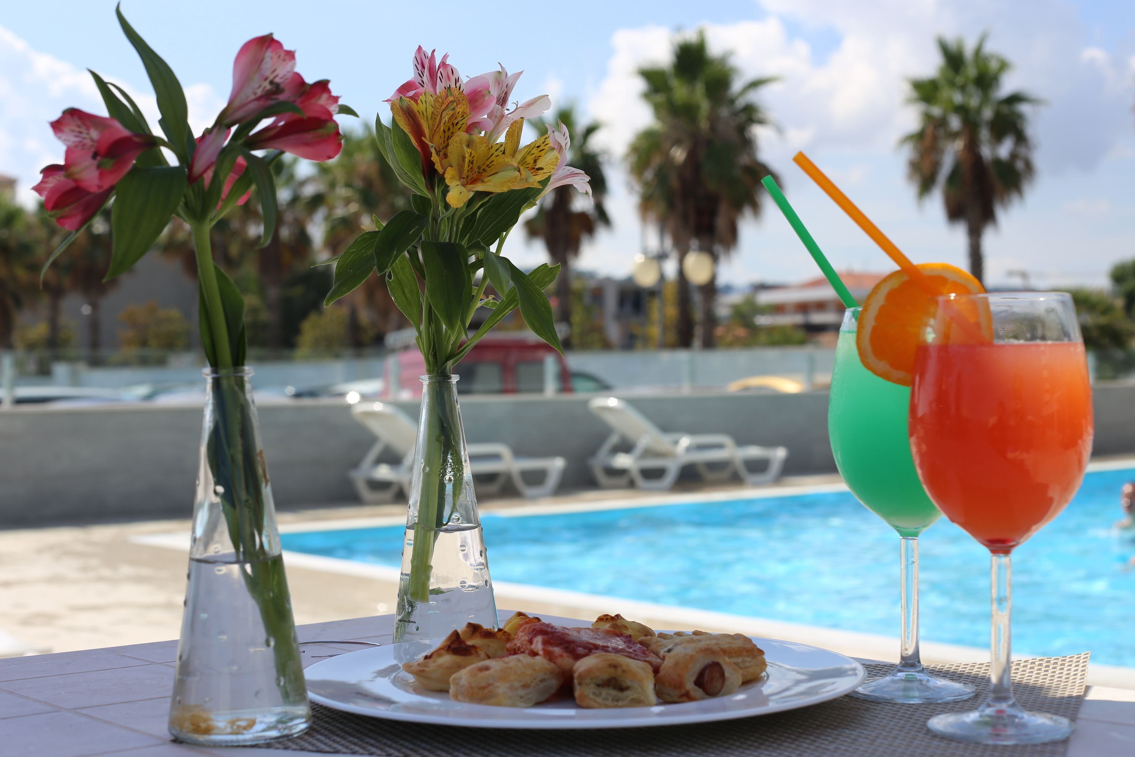 Uma mesa com um prato de comida, um copo de bebida e dois vasos de flores à beira da piscina.