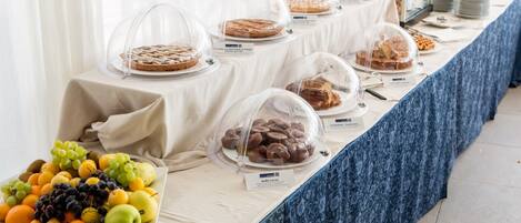 Una mesa de buffet con diversos alimentos cubiertos por campanas de cristal, entre ellos pasteles, muffins y bollería, y una selección de frutas como uvas y manzanas a un lado.