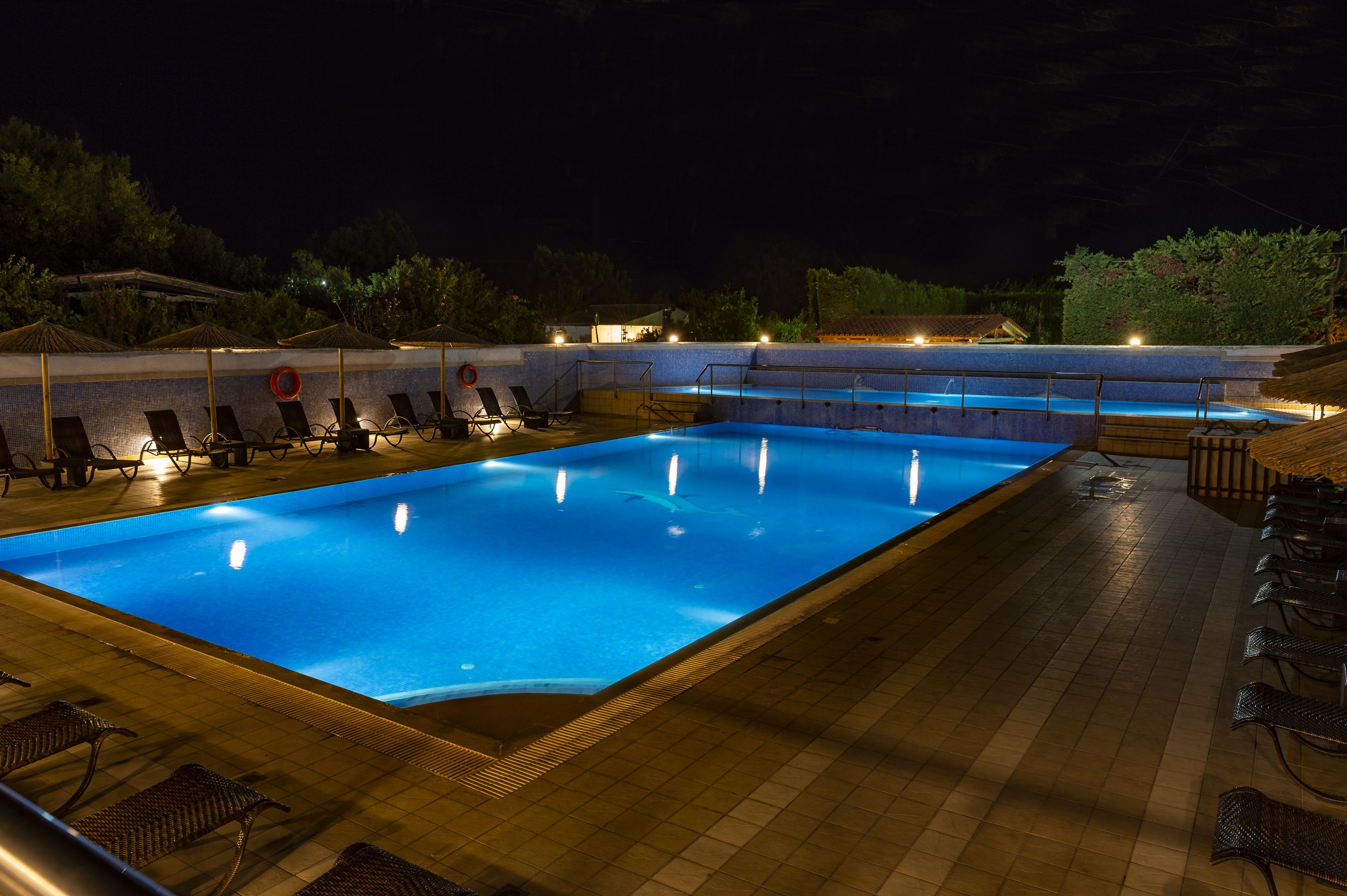 A swimming pool with lounge chairs and umbrellas, surrounded by a tiled deck and trees.
