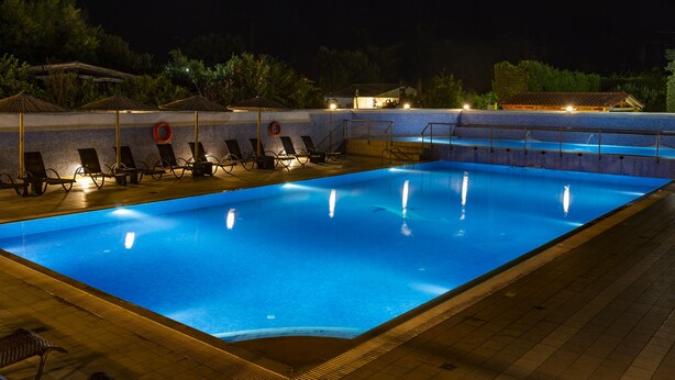 A nighttime swimming pool with lounge chairs and umbrellas, surrounded by a tiled deck and trees.