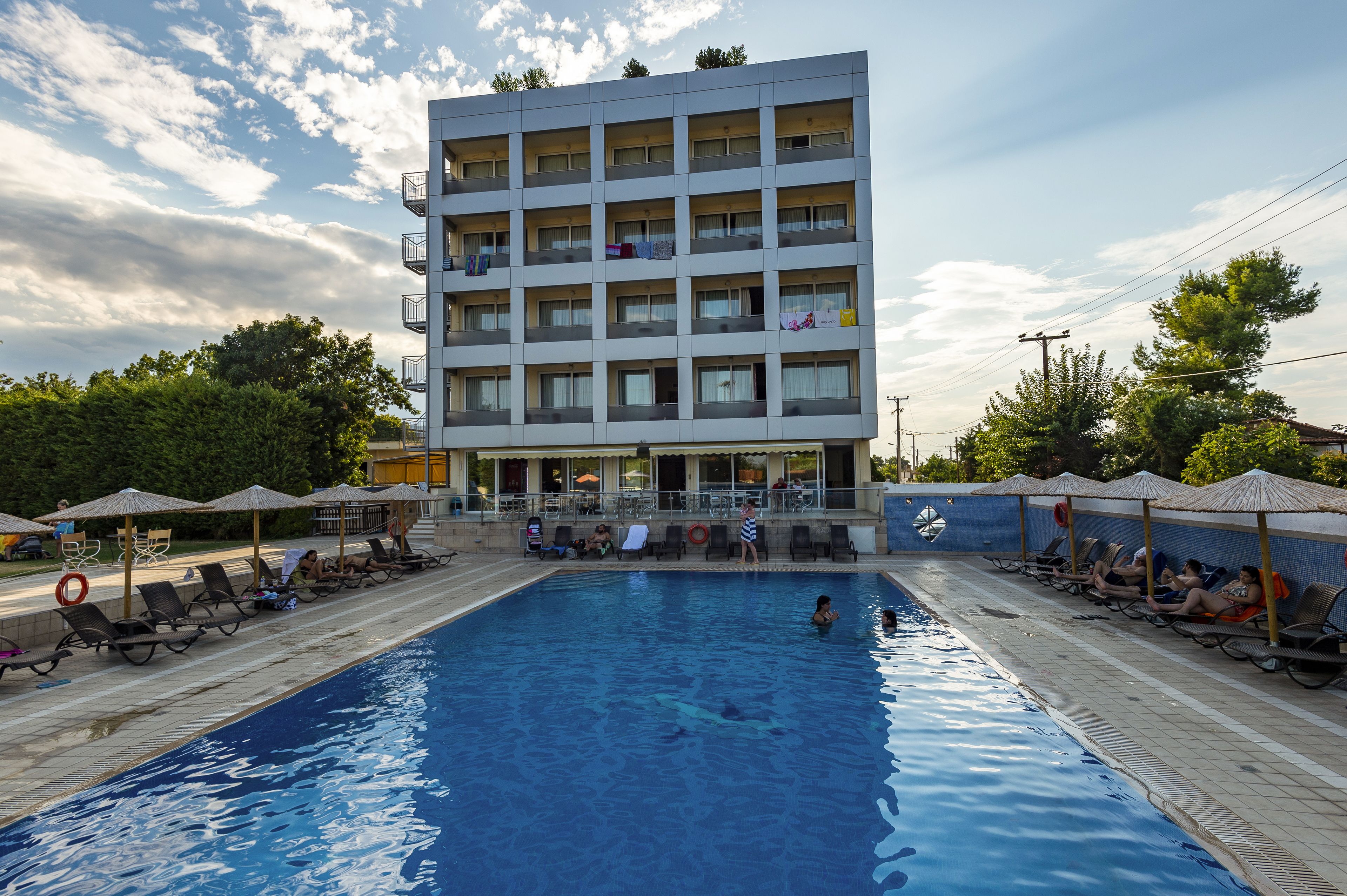 Un área de piscina con sillones y sombrillas, un edificio con balcones y un cielo despejado.