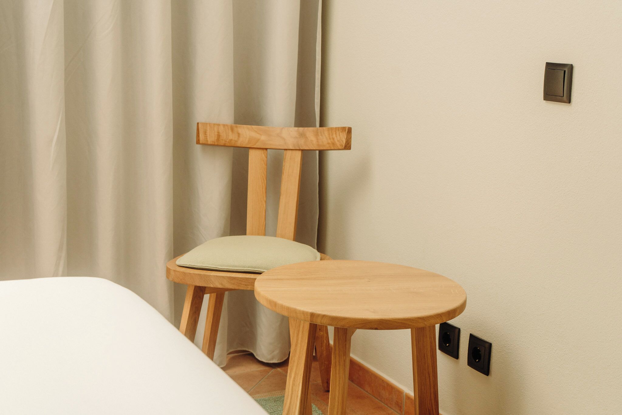 Quarto com uma cama, uma cadeira de madeira com almofada, uma pequena mesa de madeira e um interruptor de luz fixado na parede.